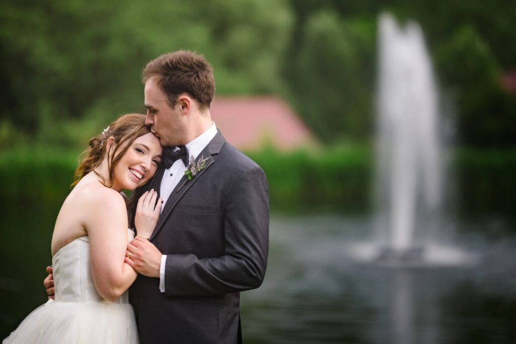Groom kissing bride's forehead while she smiles, embracing outdoors by a fountain. &mdash; Wedding at Glasbern Inn, Fogelsville, PA