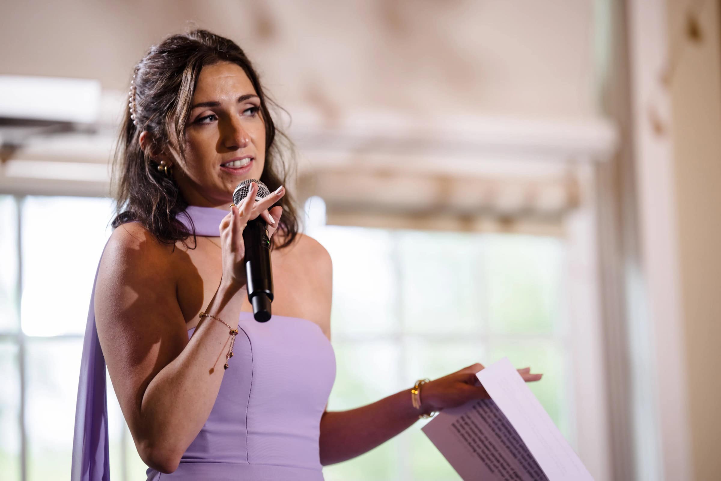Woman in lavender dress speaking into microphone at an indoor event, holding papers. — Wedding at Glasbern Inn, Fogelsville, PA