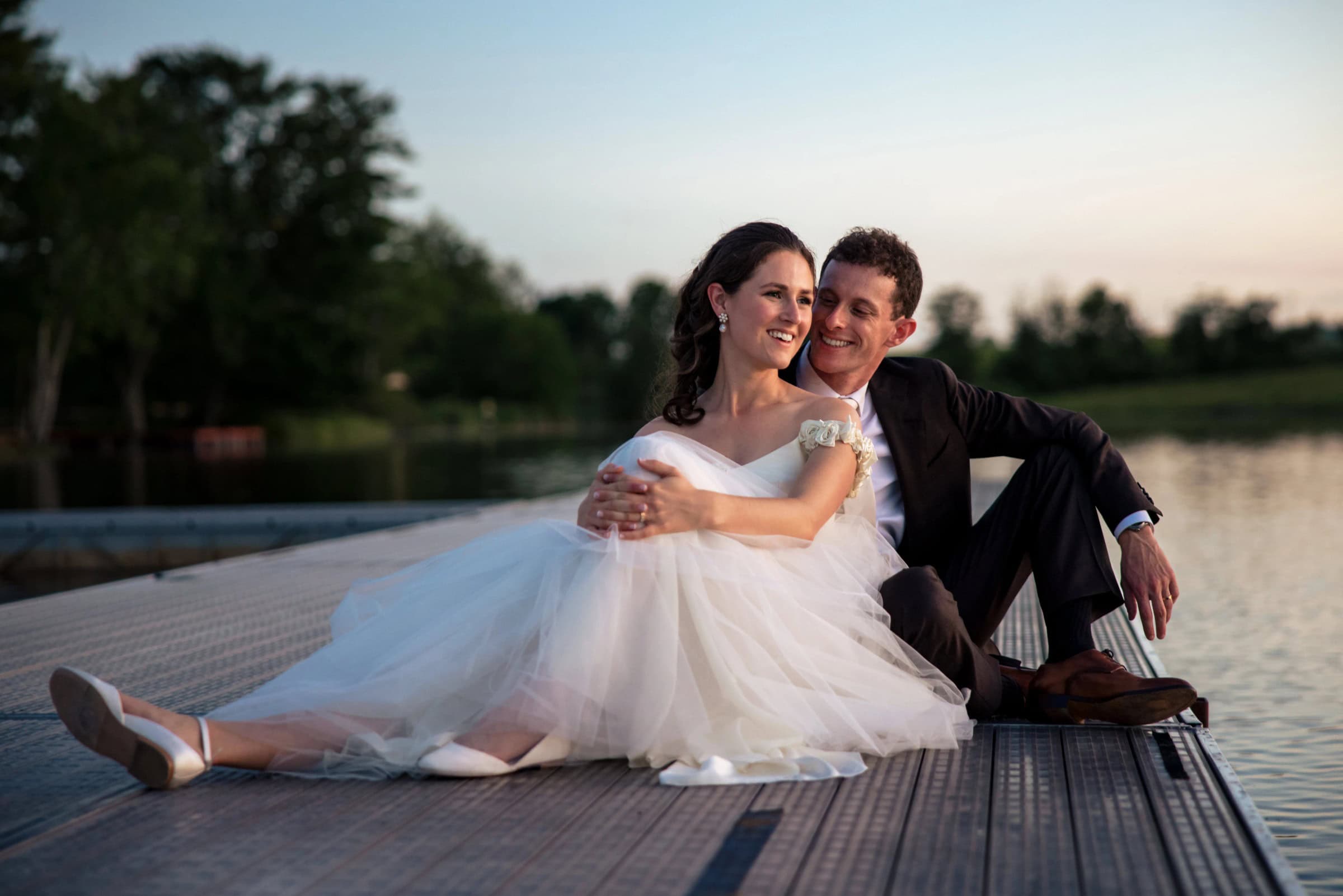 Bride and groom sitting together on a wooden deck beside water at dusk, embracing and gazing toward the horizon. &mdash; Wedding at Tyler Hill Camp, Tyler Hill, PA