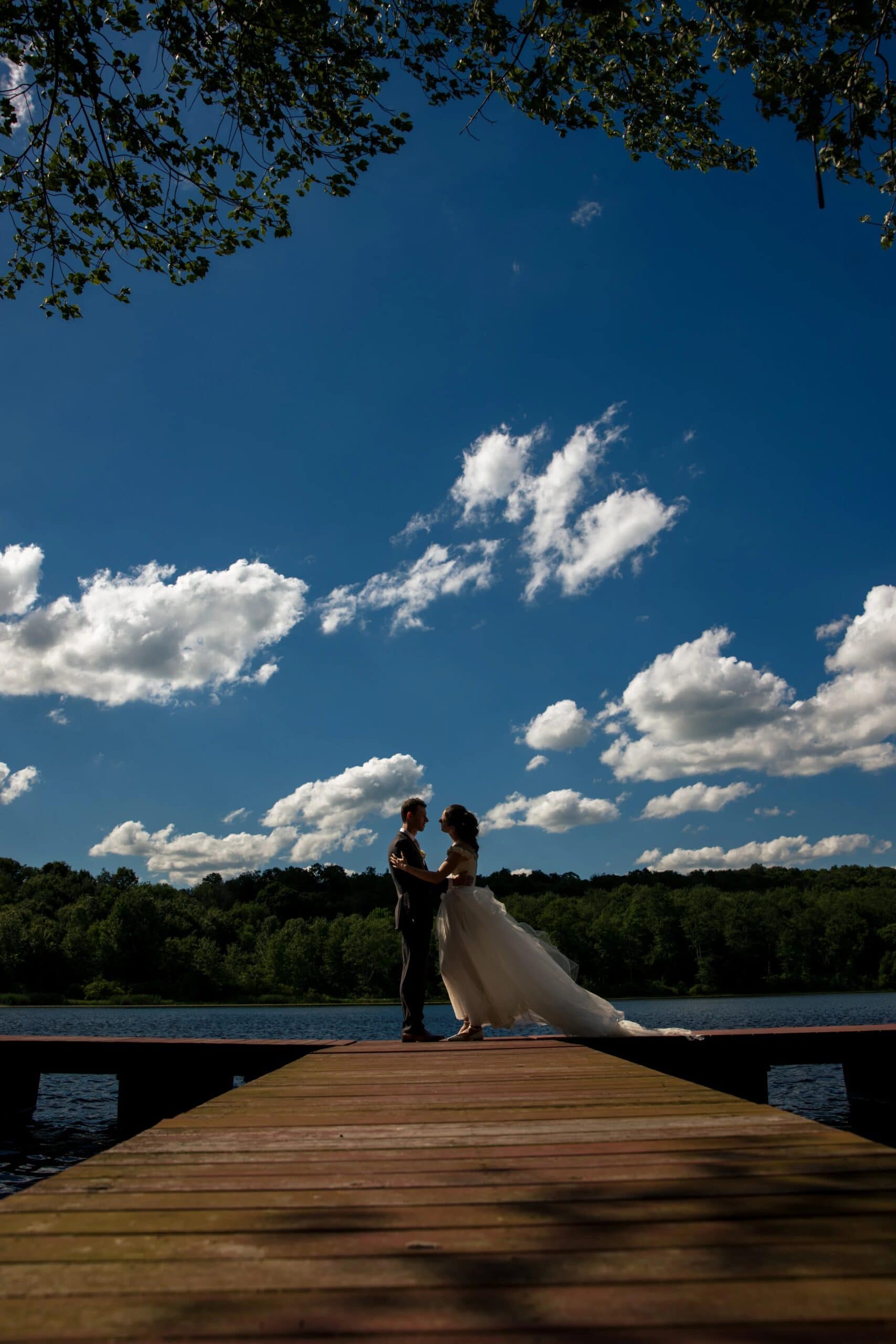 Bride and groom standing together on a wooden dock overlooking water, bride's train flowing behind her. — Wedding at Tyler Hill Camp, Tyler Hill, PA