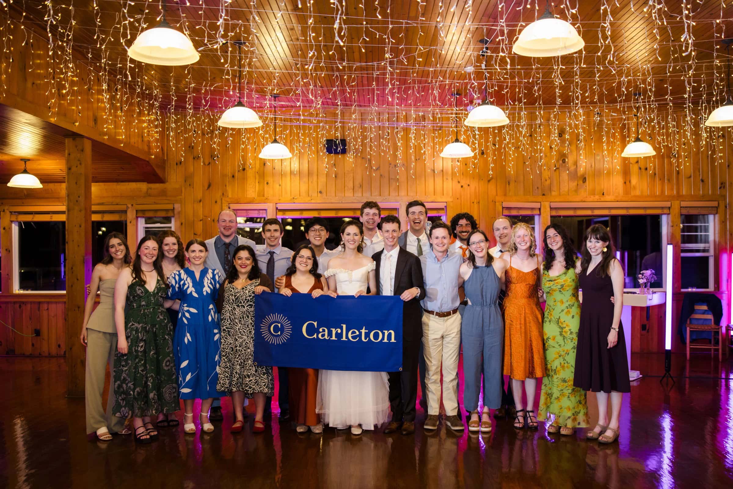 Large group holding a Carleton banner poses formally under decorative gold curtains and pendant lights. — Wedding at Tyler Hill Camp, Tyler Hill, PA