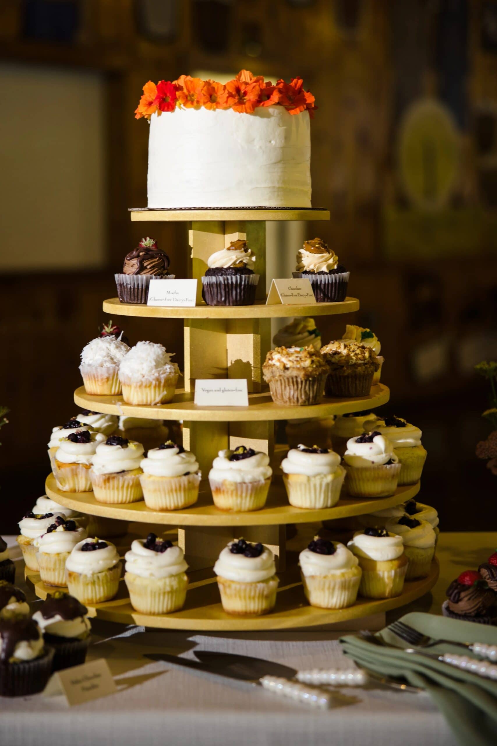 A tiered wedding cake display with cupcakes arranged on gold stands, topped with orange flowers. — Wedding at Tyler Hill Camp, Tyler Hill, PA