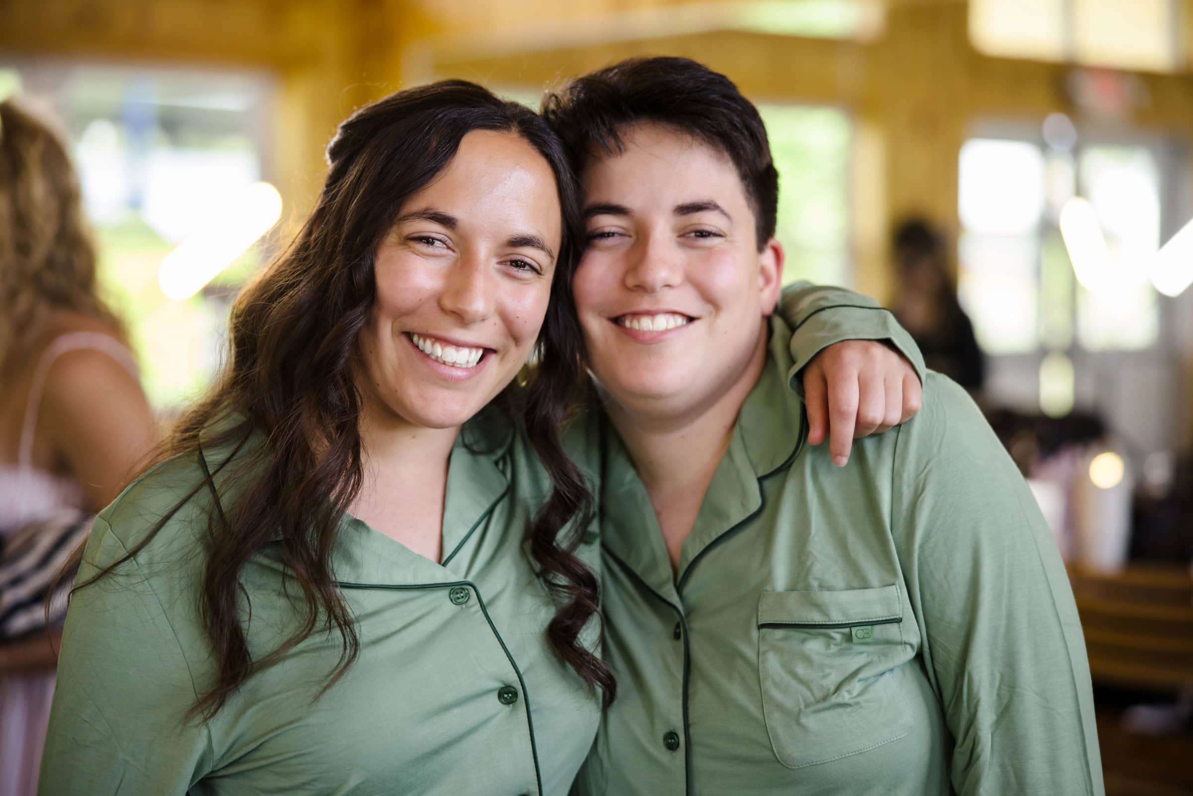 Two women in matching sage shirts smile together with arms around each other's shoulders. — Wedding at Tyler Hill Camp, Tyler Hill, PA