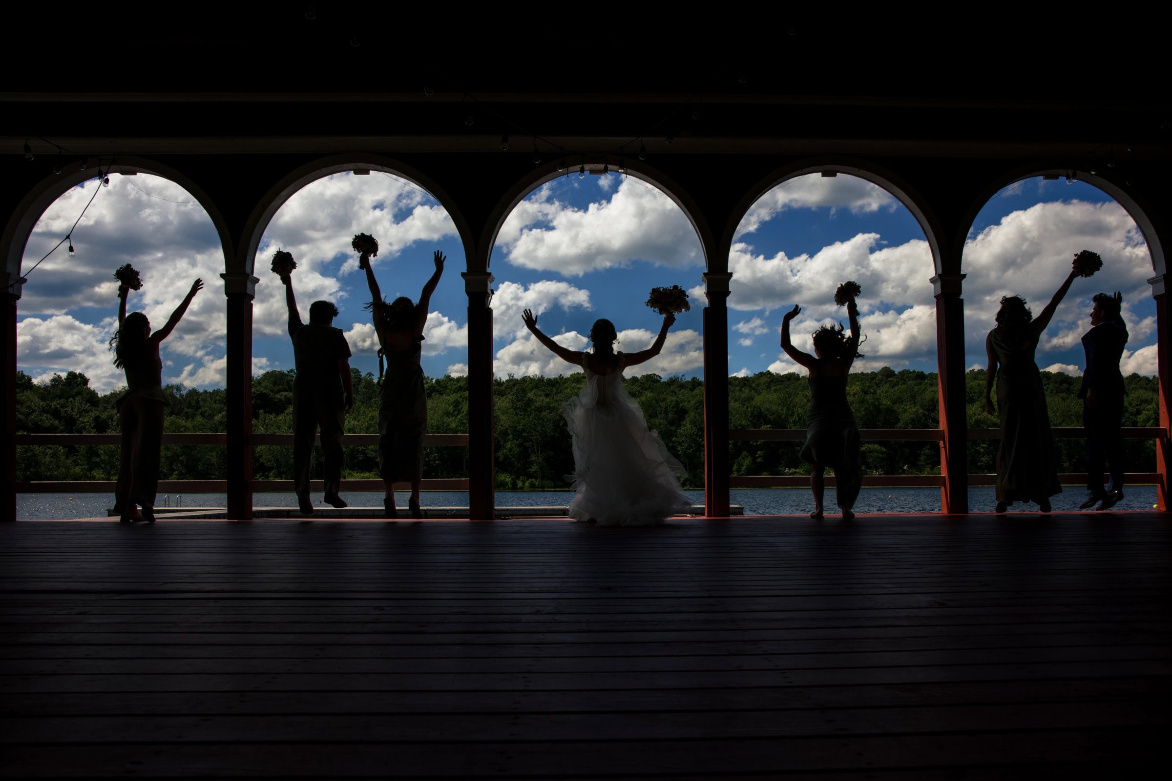 Wedding party members celebrating with raised arms, silhouetted against arched openings framing sky and landscape. — Wedding at Tyler Hill Camp, Tyler Hill, PA