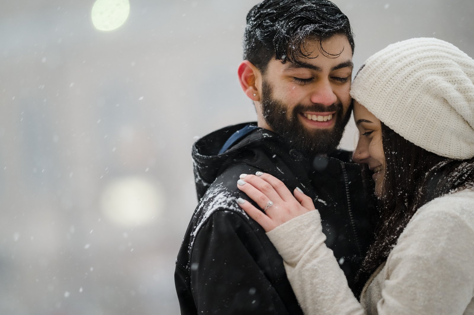 Couple cuddling close as snow settles on their hair and winter clothing.