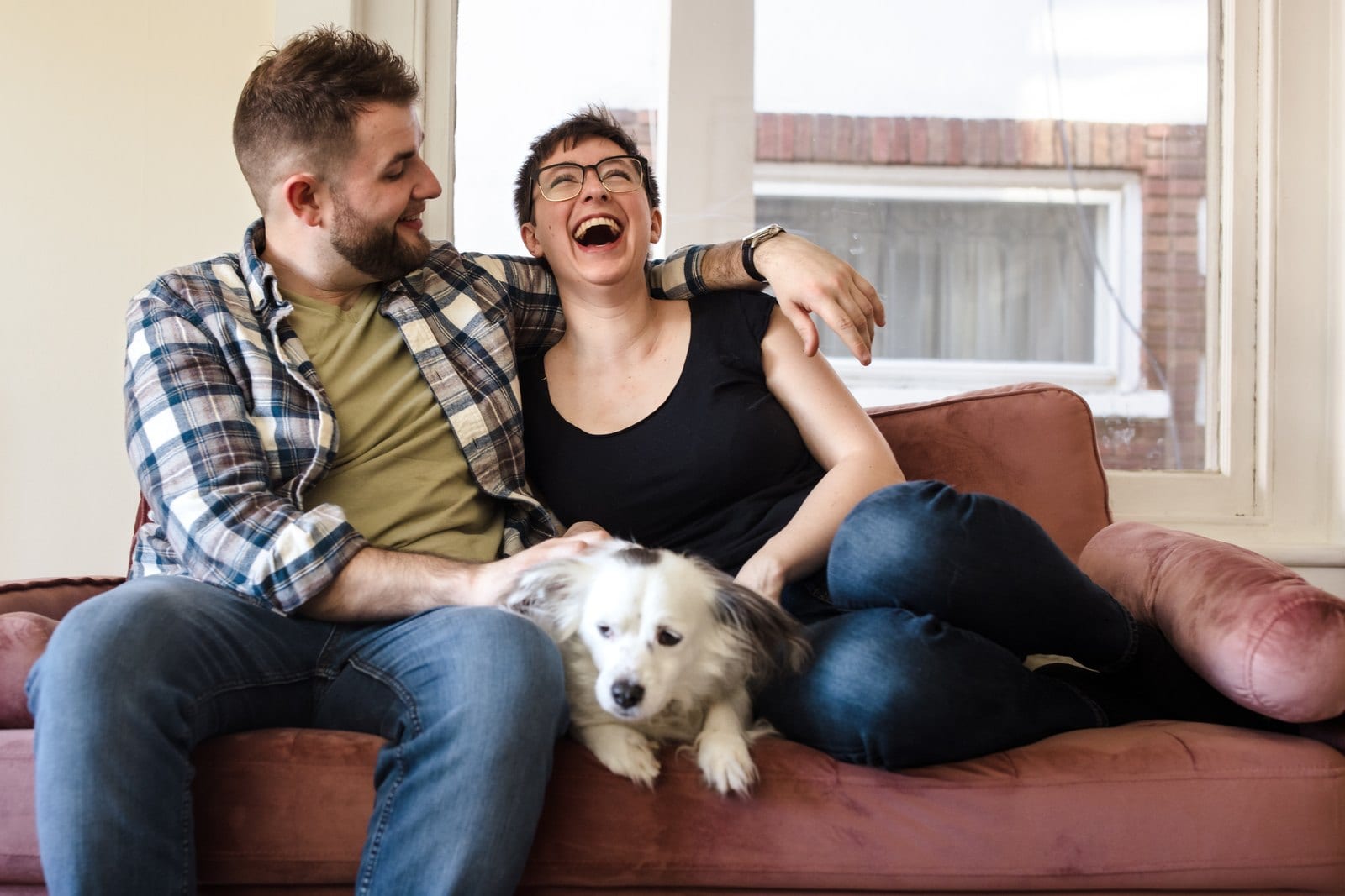 Couple laughing together on a sofa with their dog stretched across their laps.