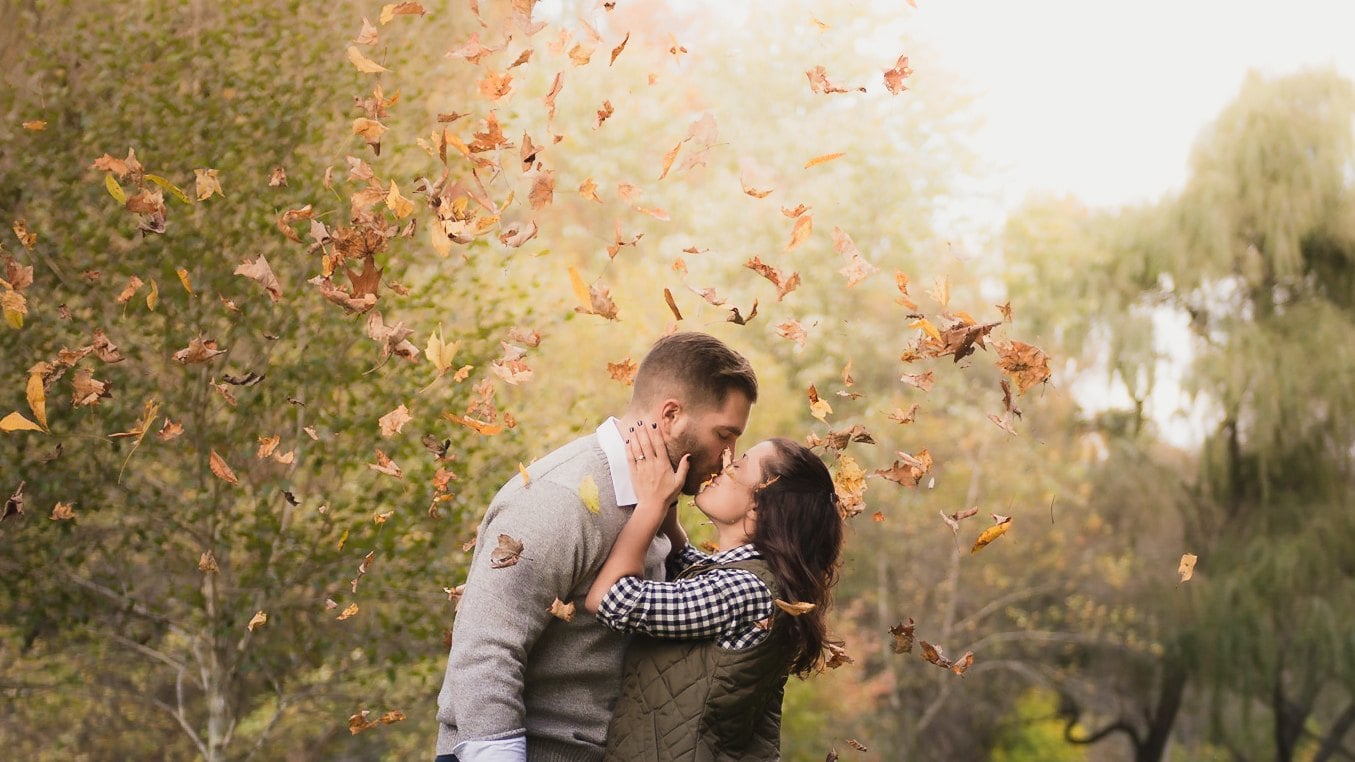 Couple kissing beneath falling autumn leaves in warm afternoon light.