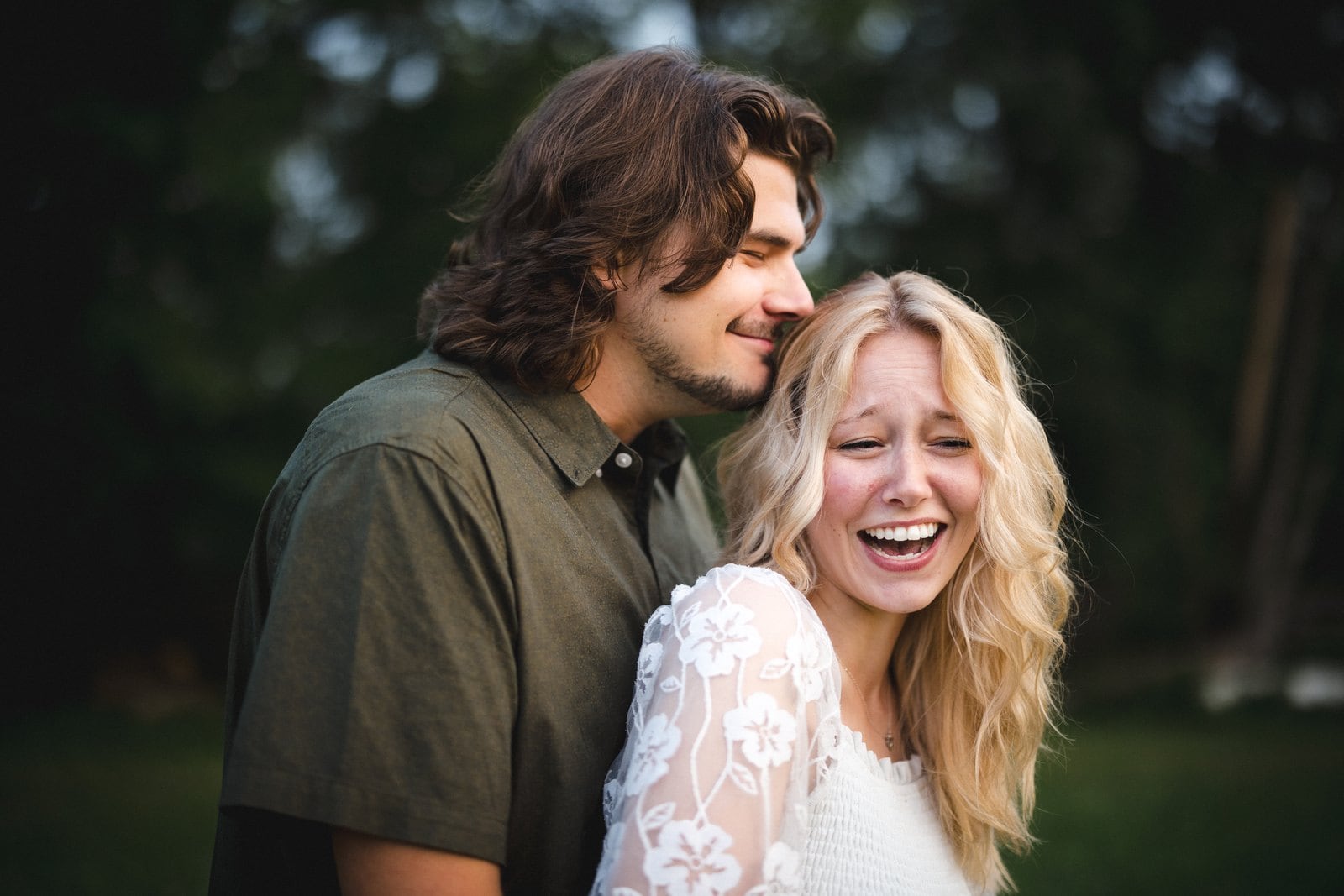 Woman laughing as her partner kisses her cheek against a soft green background.