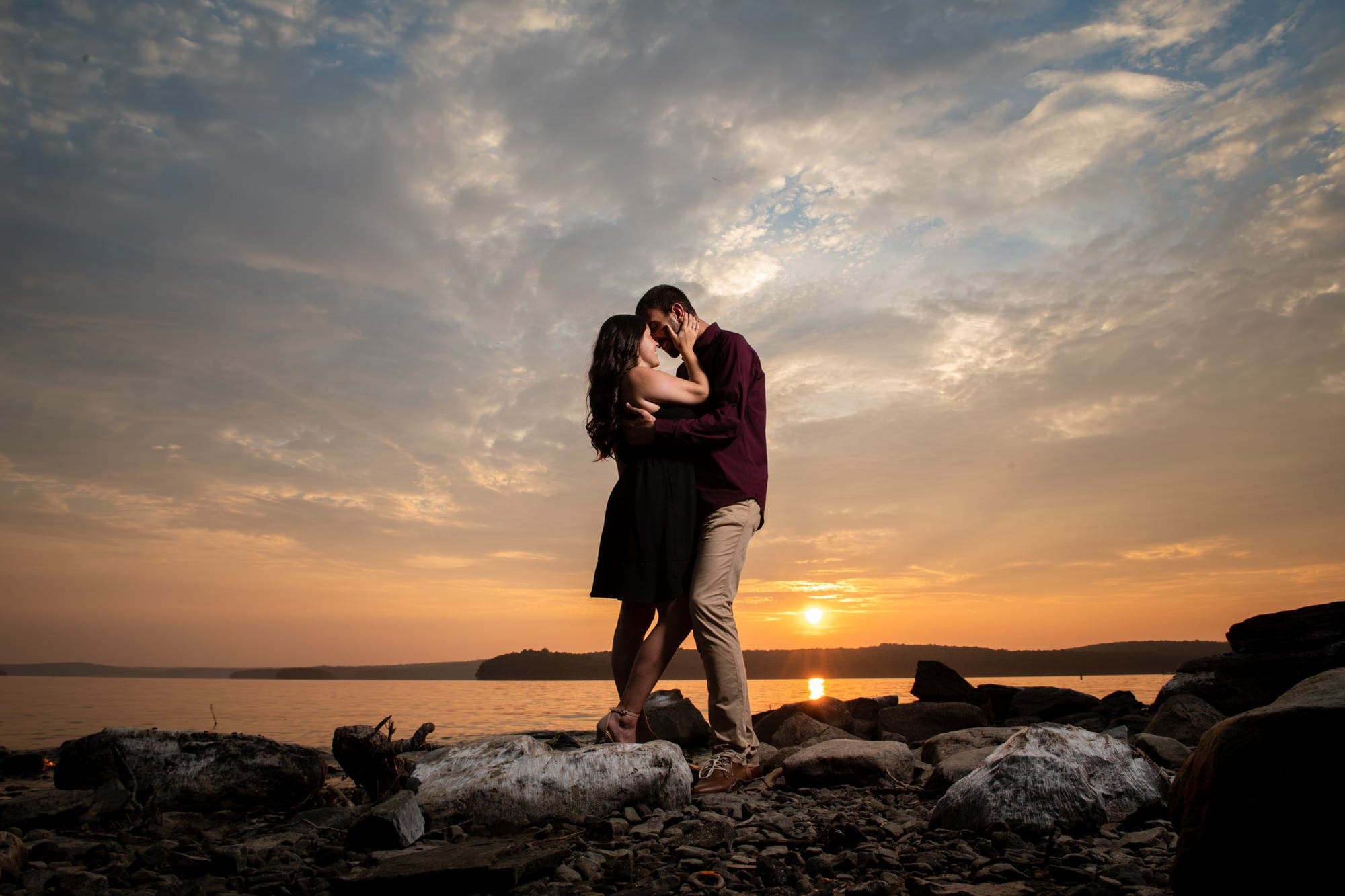 Couple embracing on a rocky lakeshore beneath an orange sunset and sweeping clouds.
