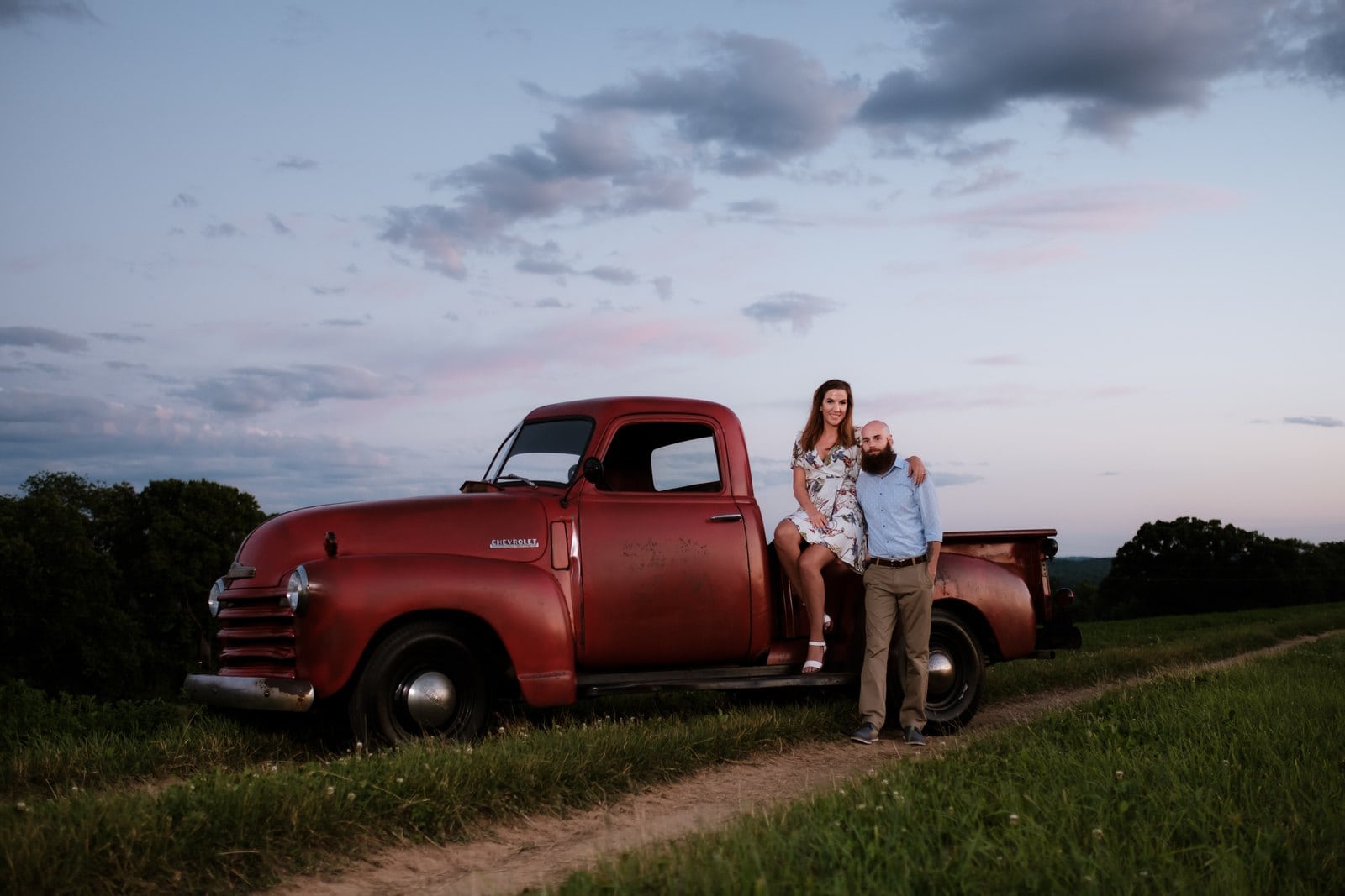 Couple leaning together beside a vintage red pickup truck under a pink and blue evening sky.