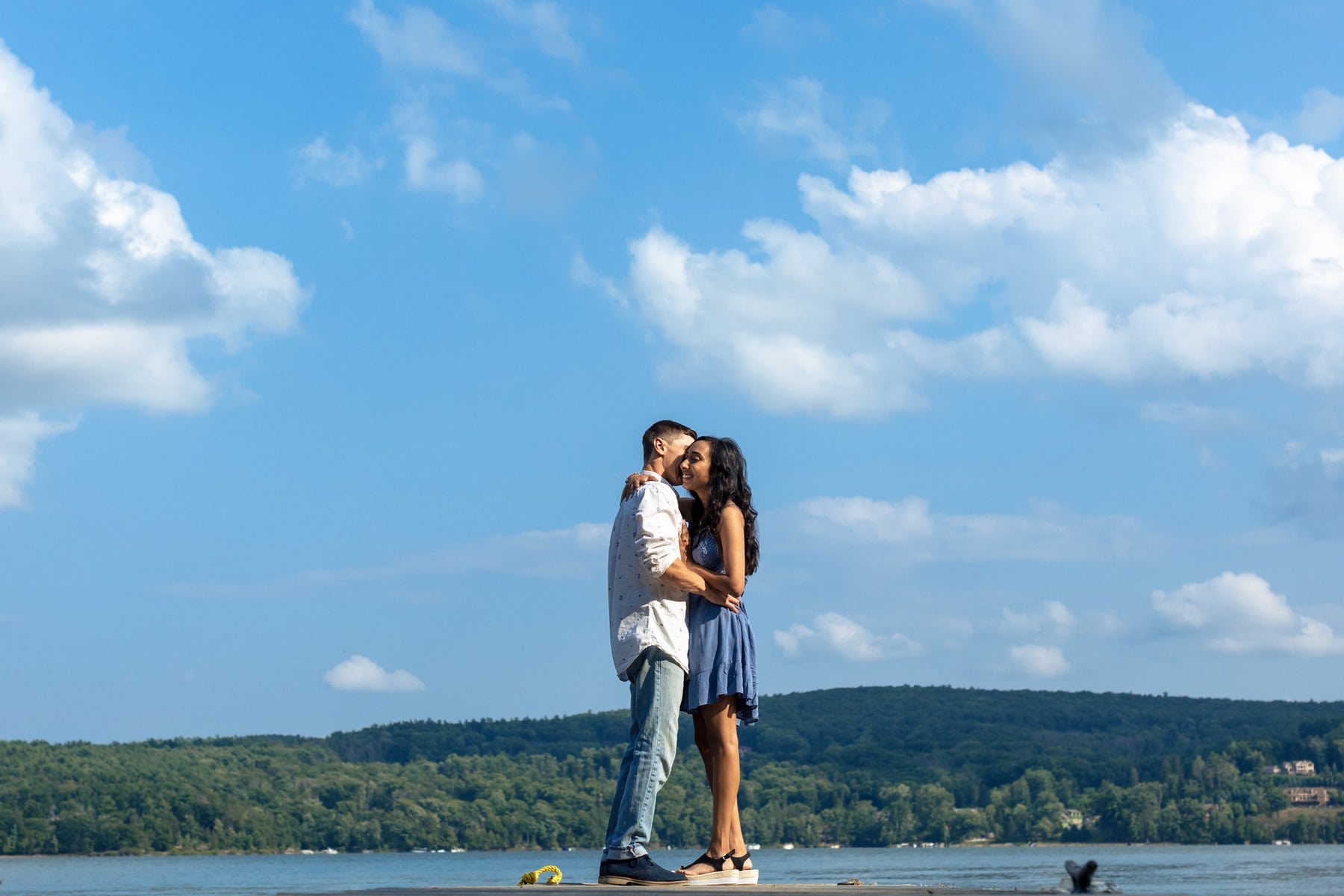 Couple kissing beside Lake Wallenpaupack beneath a wide blue sky with scattered clouds.