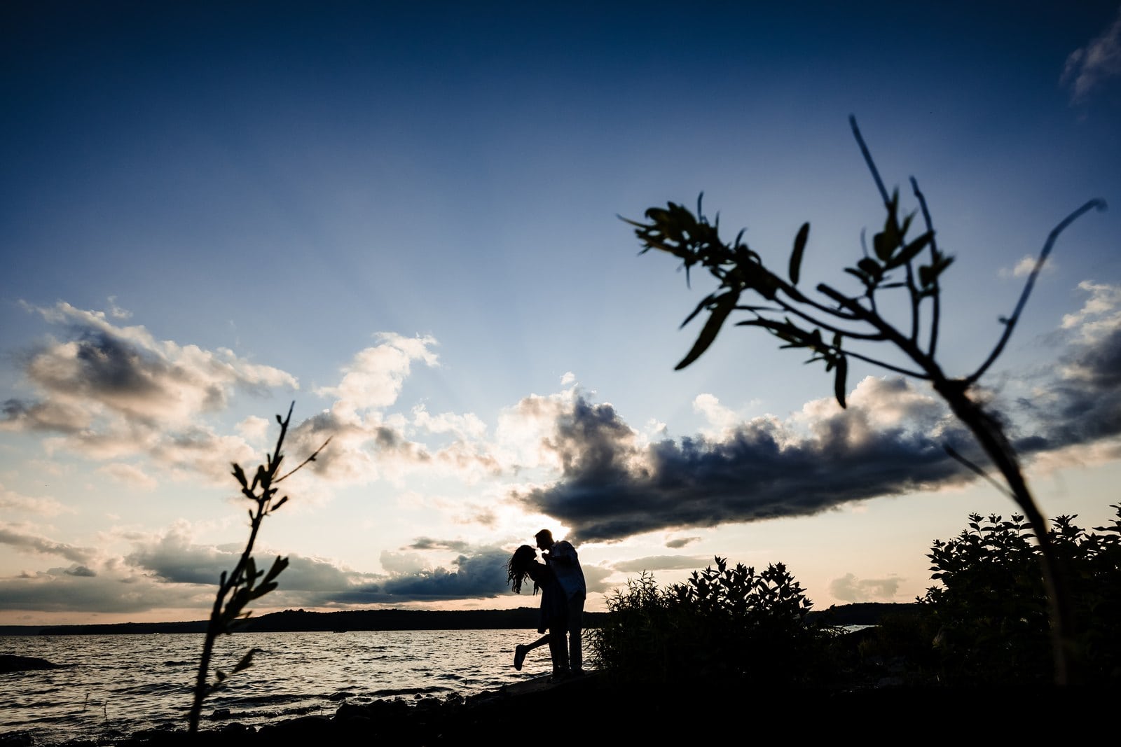 Silhouetted couple embracing beside Lake Wallenpaupack beneath dramatic clouds and overhanging branches.